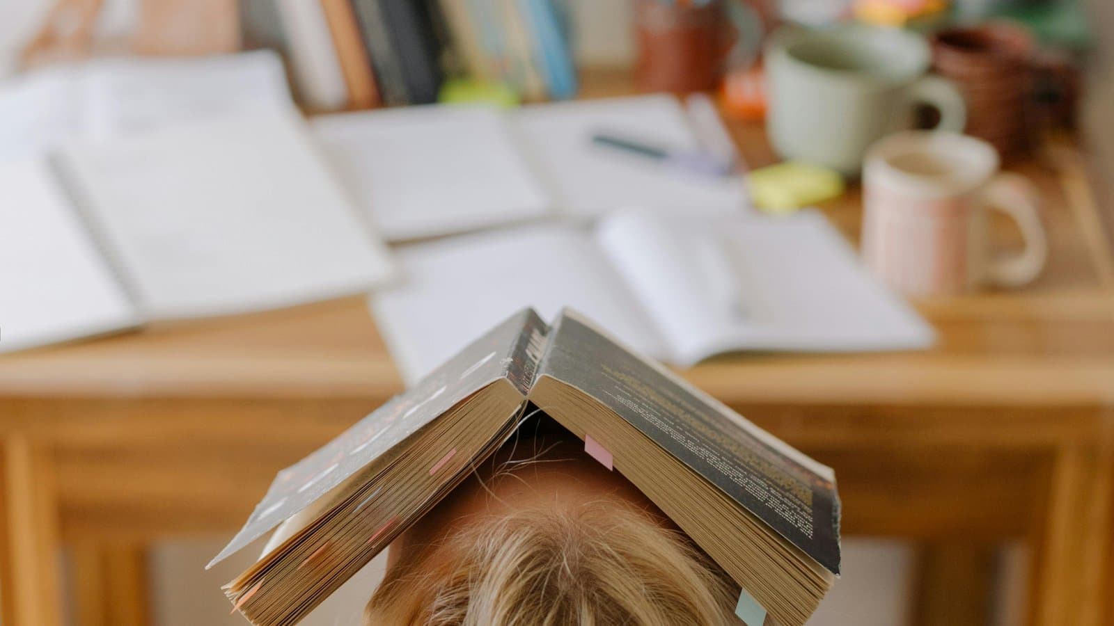 Erschöpfte Person mit aufgeschlagenem Buch über dem Kopf am vollen Schreibtisch, Sinnbild für die Frage, ob das Uni-Rep ausreicht