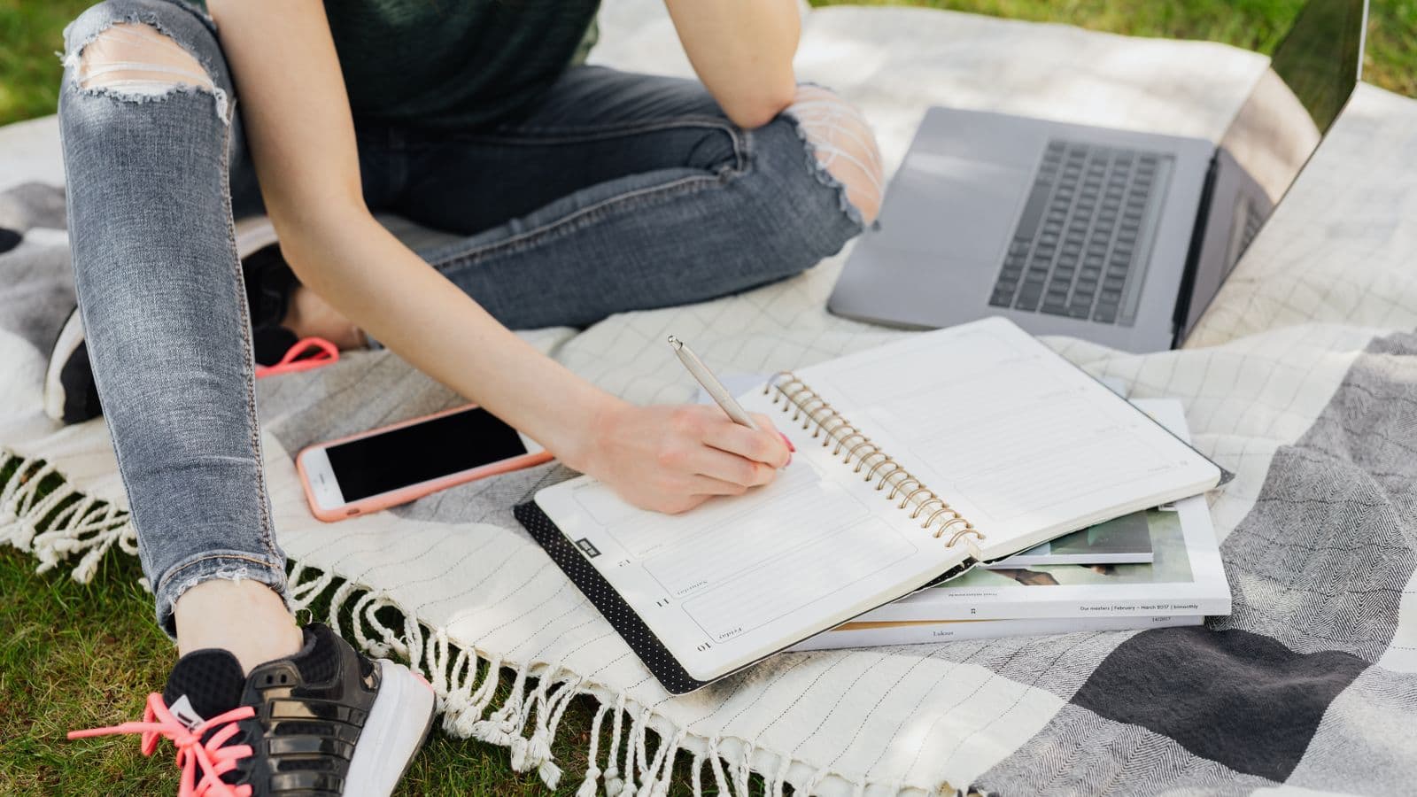 Studentin schreibt im Park auf einer Decke ihren Wochenplan in einen Planer, daneben Laptop und Smartphone.
