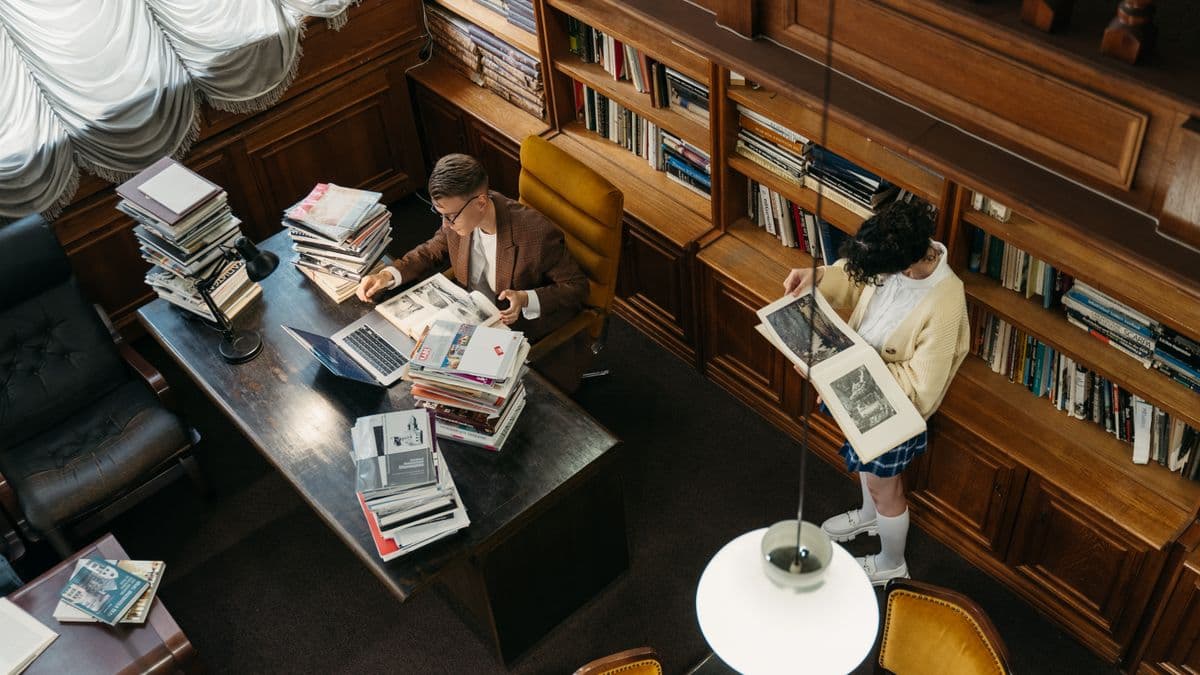 Zwei Menschen studieren in einer holzvertäfelten Bibliothek, eine Person liest an einem Schreibtisch mit Bücherstapeln, eine weitere blättert an einem Bücherregal.