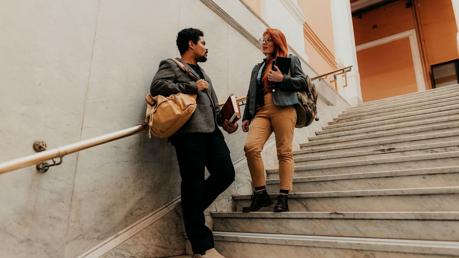 Zwei Jura-Studierende unterhalten sich mit Büchern und Rucksäcken auf einer Treppe vor einem Universitätsgebäude.