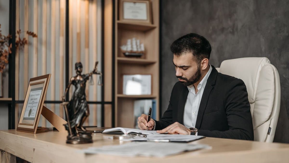 Ein Anwalt im Anzug arbeitet an Dokumenten an seinem Schreibtisch, daneben eine Justitia-Figur.