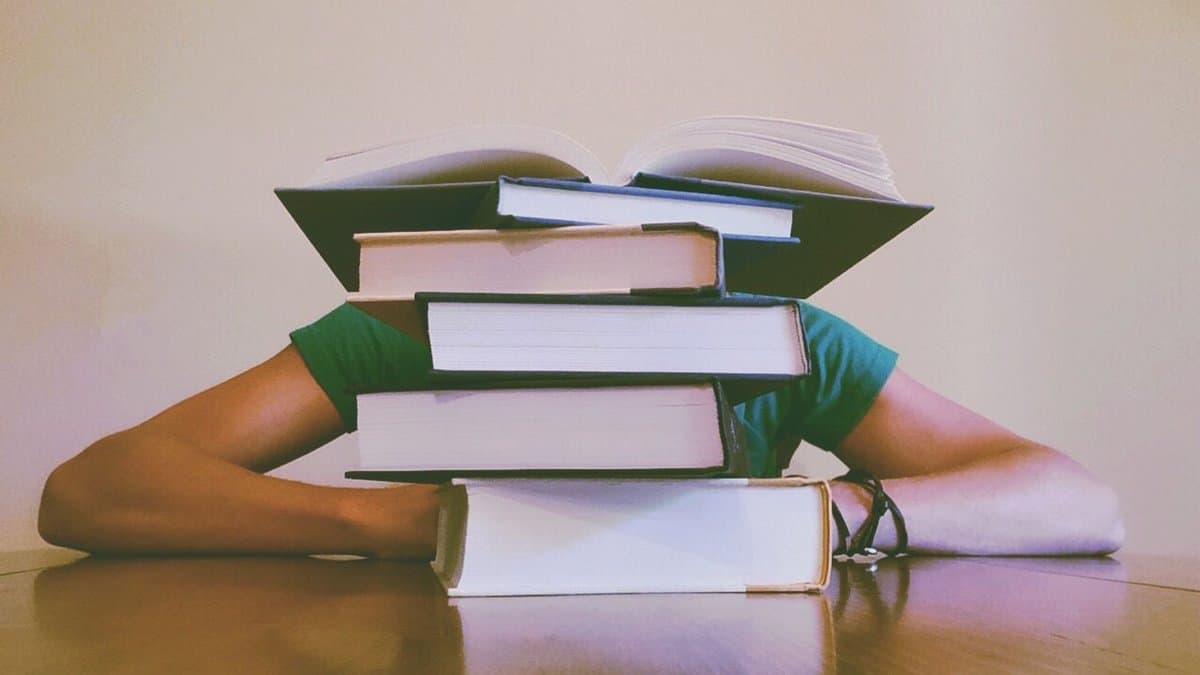Studierende Person sitzt fast verdeckt hinter einem hohen Bücherstapel am Schreibtisch