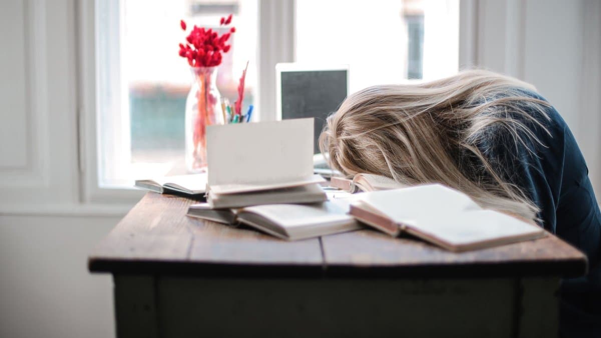 Studentin sitzt erschöpft über aufgeschlagenen Büchern an einem Schreibtisch und stützt den Kopf ab.