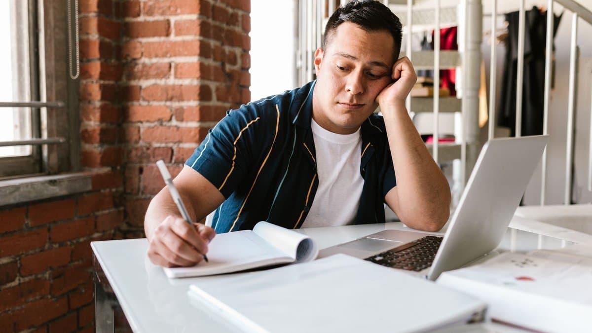 Ein Student sitzt gelangweilt mit Laptop und Notizbuch am Schreibtisch.