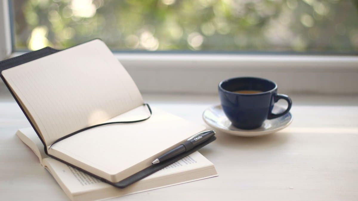 Aufgeschlagenes Notizbuch mit einer Tasse Kaffee auf einem Schreibtisch im Morgenlicht am Fenster.