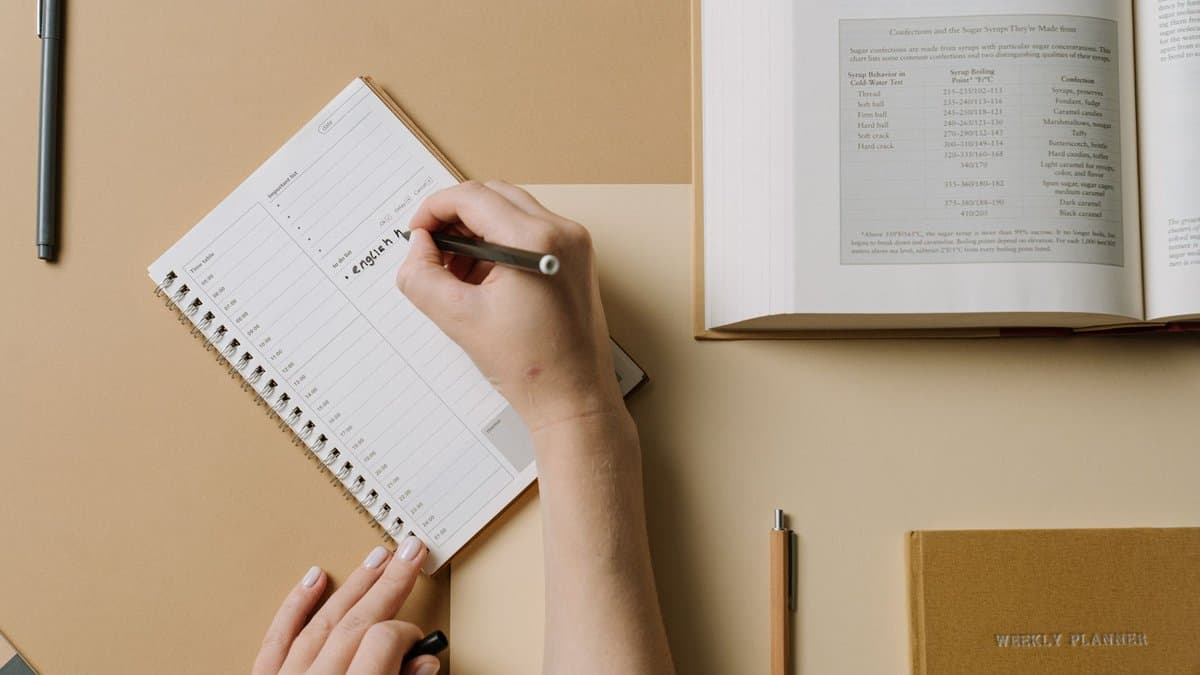 Hände tragen einen Wochenplan in einen Kalender ein, daneben liegt ein aufgeschlagenes Lehrbuch