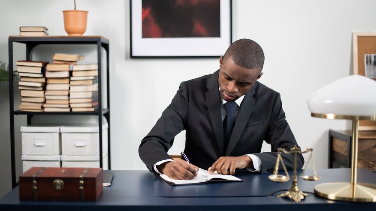 Person im Anzug schreibt handschriftlich an einem Schreibtisch mit aufgeschlagenem Buch, einer Justitia-Waage und Gesetzbüchern im Regal