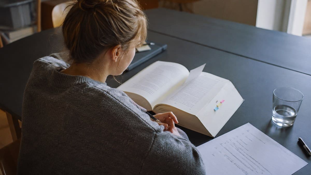 Studentin sitzt am dunklen Tisch, liest in einem dicken Gesetzbuch mit farbigen Markern und schreibt daneben eine handschriftliche Gliederung