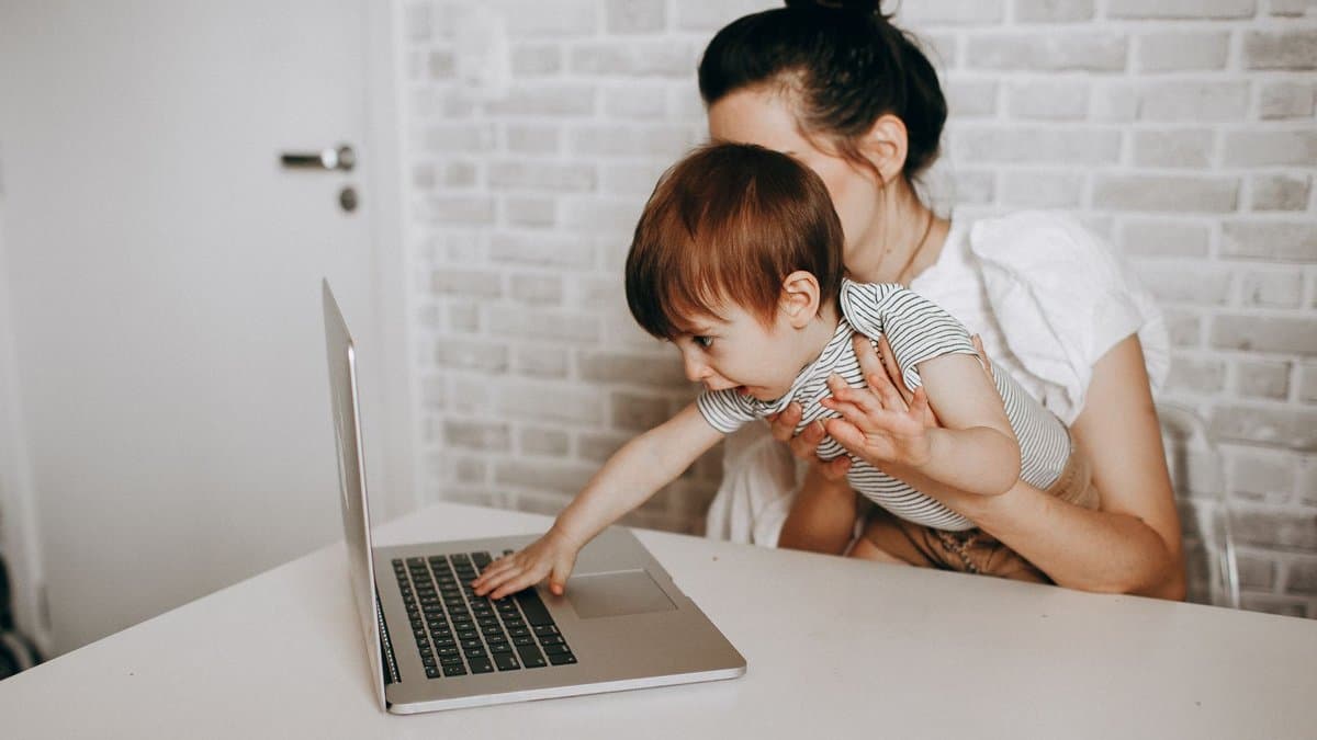 Eine Mutter hält ihr Kleinkind auf dem Arm, während sie am Laptop am Tisch arbeitet