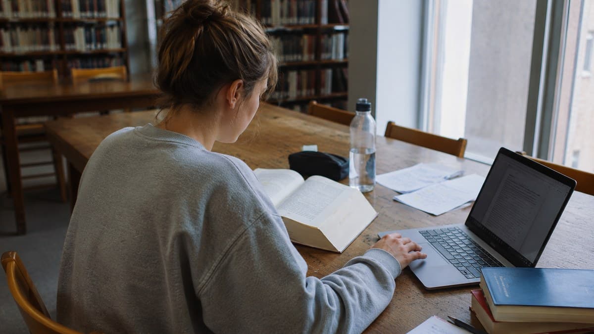 Studentin in grauem Sweatshirt liest am Bibliothekstisch in einem Buch, daneben Laptop und Notizen