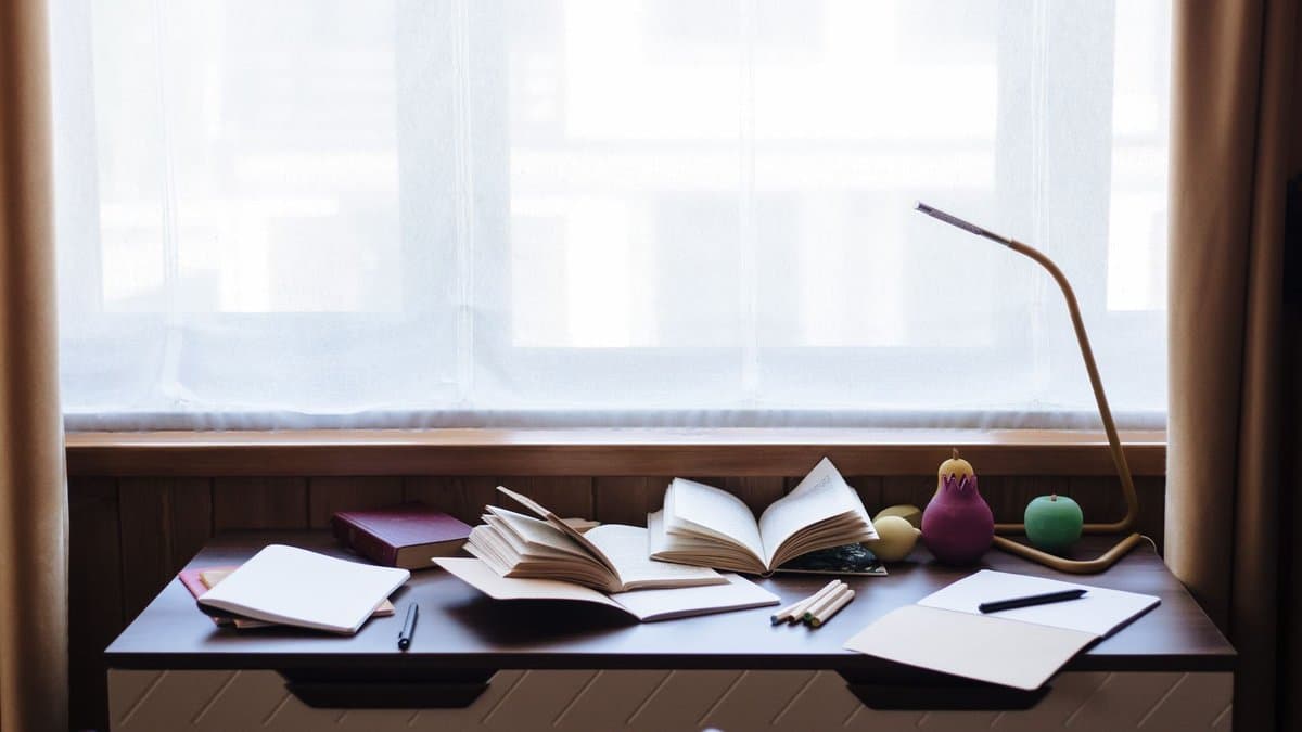 Schreibtisch am Fenster mit aufgeschlagenen Büchern, Notizheften und einer Leselampe