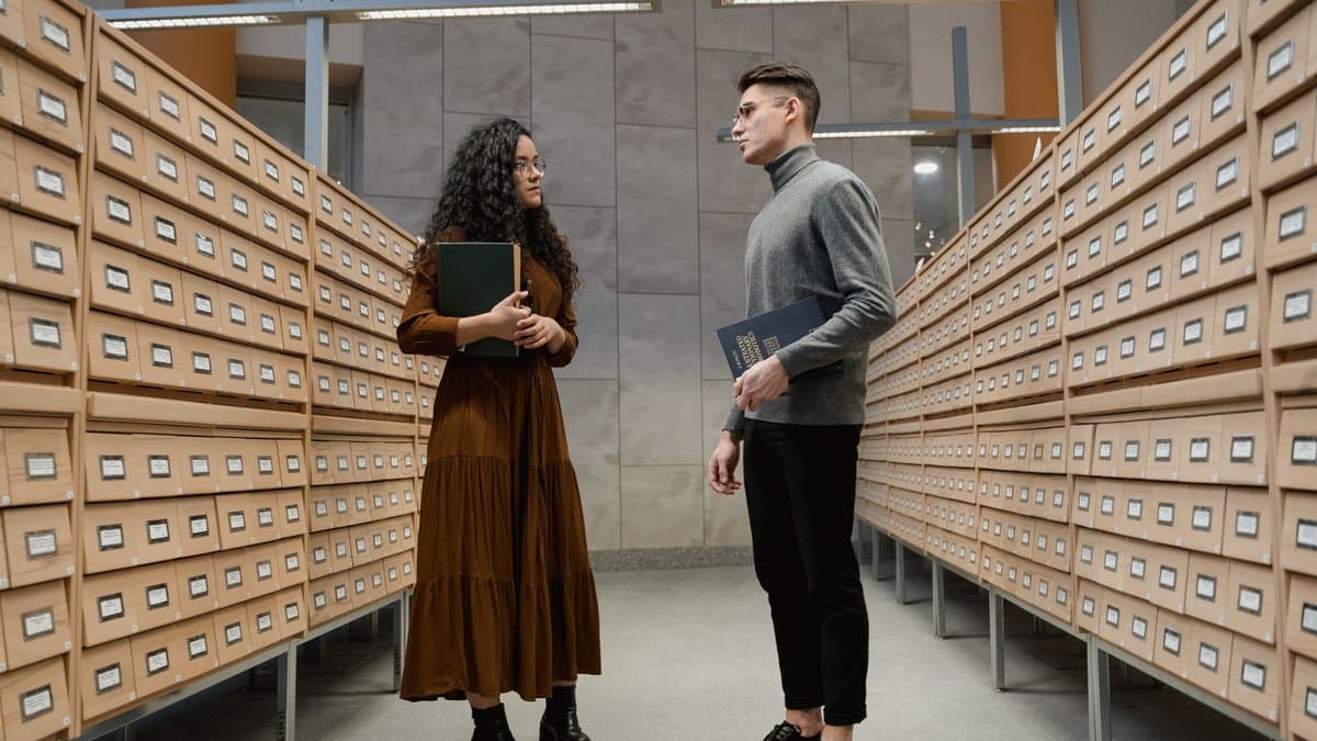 Zwei Studierende stehen im Gang einer Bibliothek und sprechen über zwei Bücher