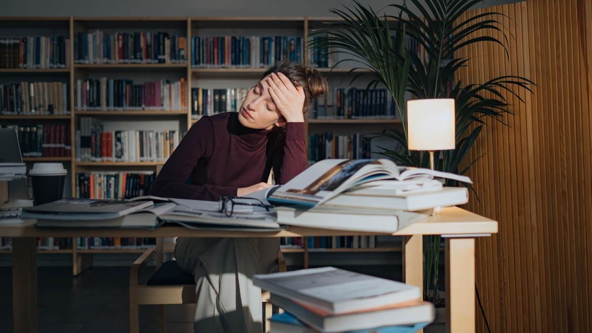 Studentin sitzt in einer Bibliothek an einem Tisch voller aufgeschlagener Bücher und Bücherstapel