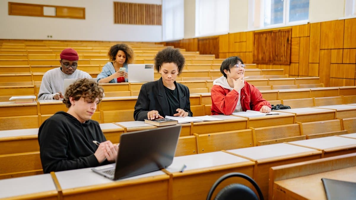 Studierende sitzen mit Laptops und Notizen in einem Hörsaal mit Holzbänken