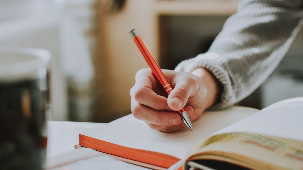 Hand mit rotem Stift über einem aufgeschlagenen Notizbuch an einem hellen Schreibtisch