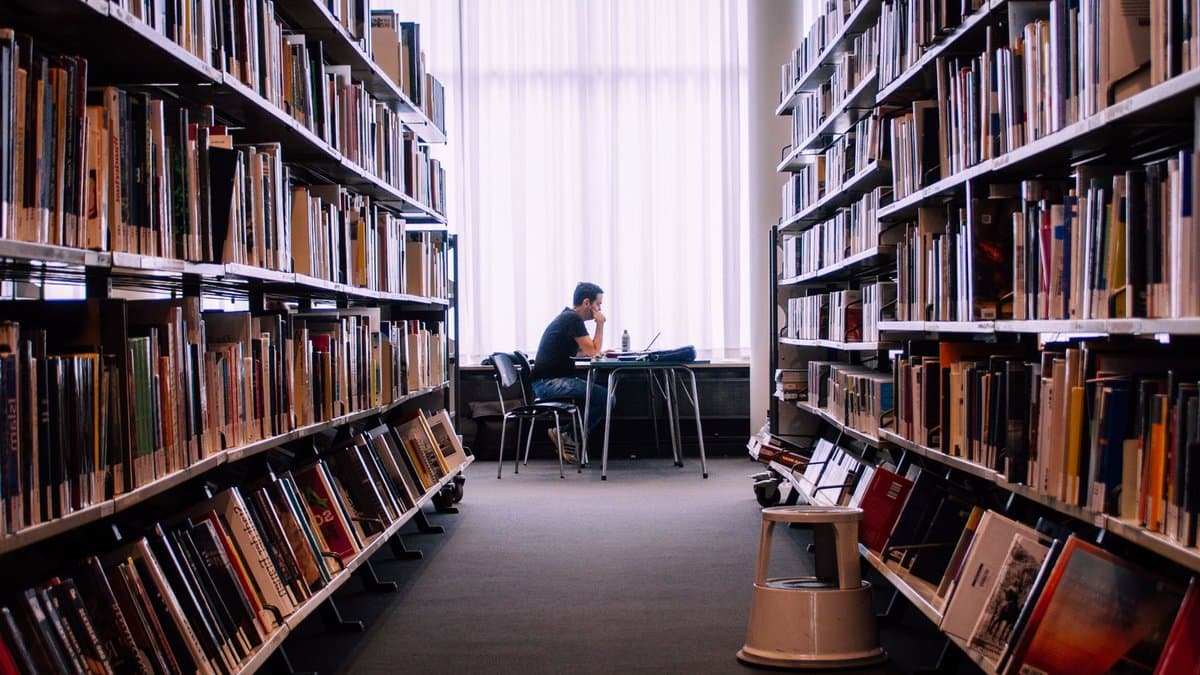 Eine Person sitzt allein an einem Tisch am Fenster einer großen Bibliothek, links und rechts hohe Bücherregale