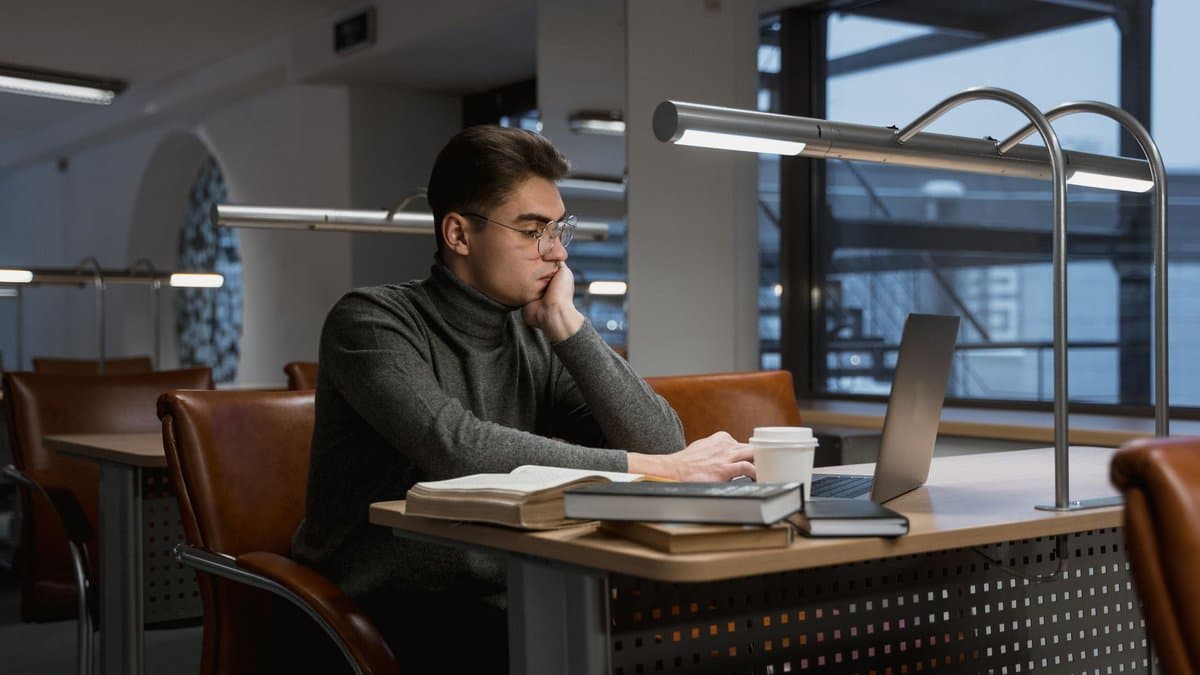 Student sitzt in einer Bibliothek am Laptop, davor aufgeschlagene Bücher und ein Kaffeebecher