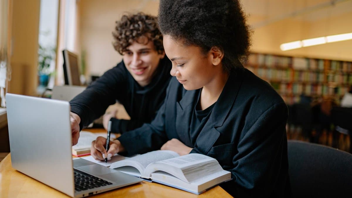 Zwei Studierende lernen gemeinsam in einer Bibliothek mit Laptop, aufgeschlagenem Buch und Notizen