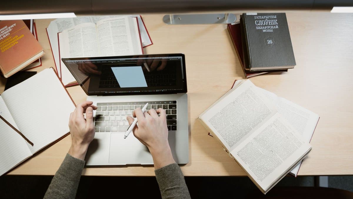 Blick von oben auf einen Schreibtisch: Hände am Laptop, daneben aufgeschlagene Bücher und ein Notizblock
