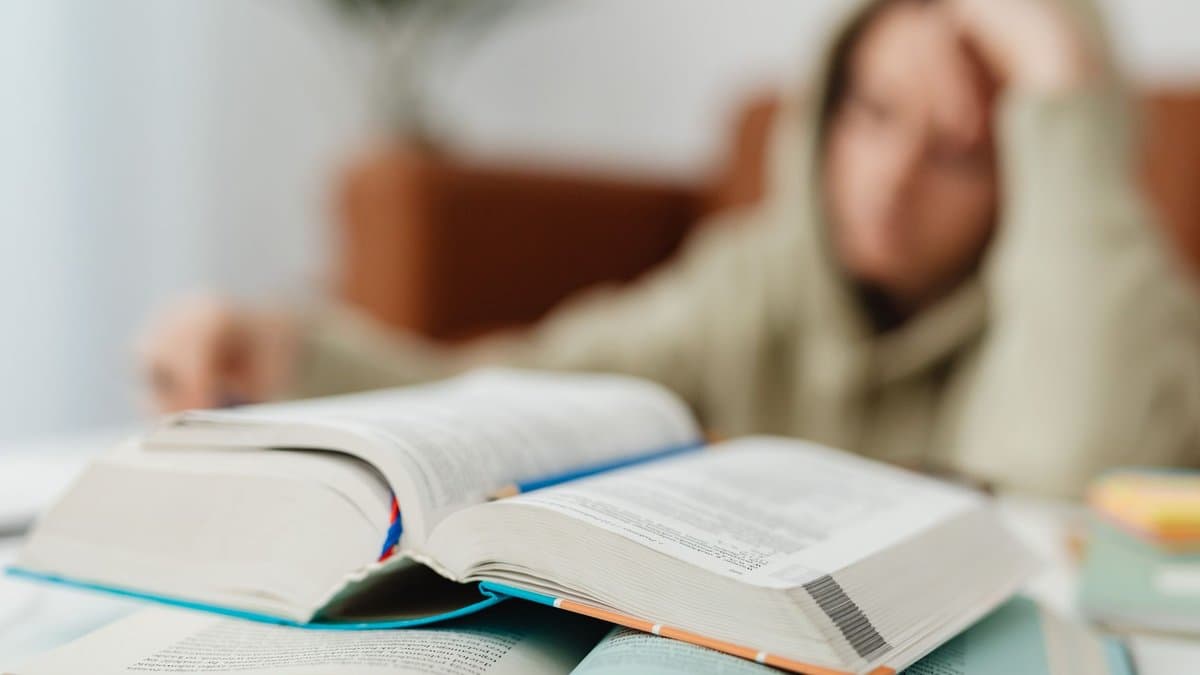 Aufgeschlagene Bücher im Vordergrund, dahinter unscharf eine erschöpfte Person am Schreibtisch