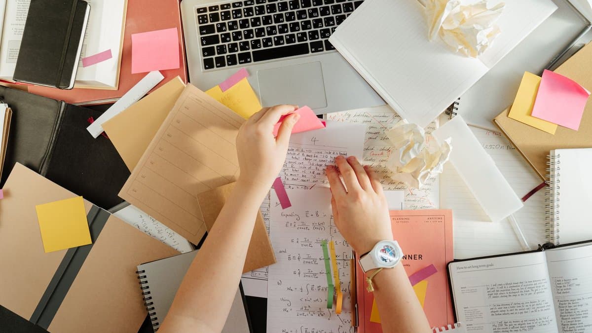 Blick von oben auf einen Schreibtisch mit handschriftlichen Notizen, Haftzetteln und Notizbüchern, zwei Hände sortieren die Zettel