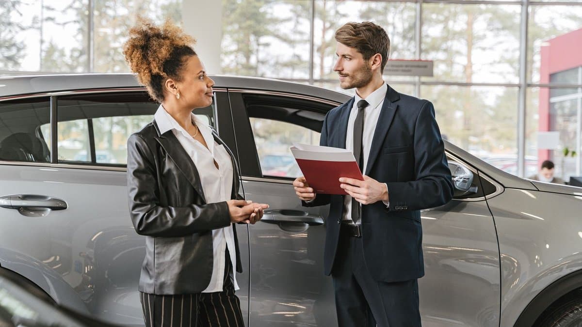 Verkäuferin und Käufer stehen mit einem Vertragsordner neben einem Auto im Autohaus