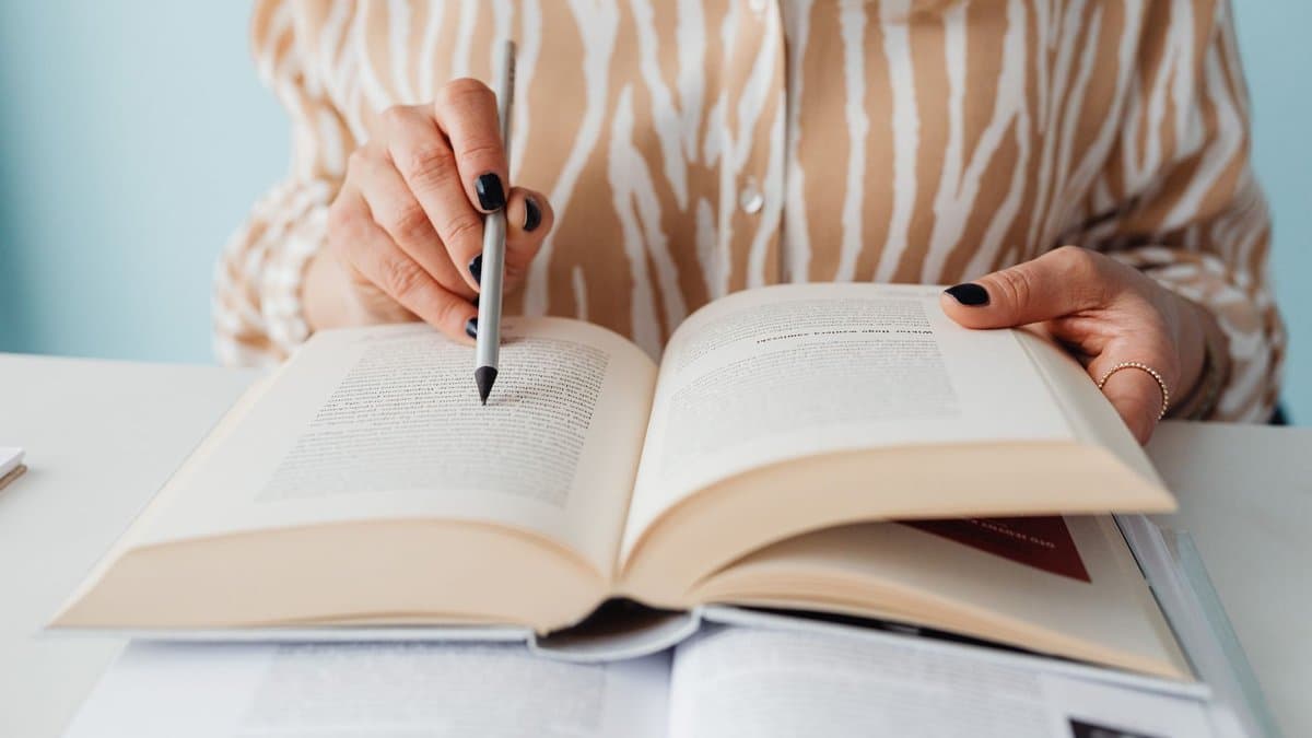 Hände, die mit einem Stift auf eine Textstelle in einem aufgeschlagenen Buch zeigen, daneben ein zweites aufgeschlagenes Buch