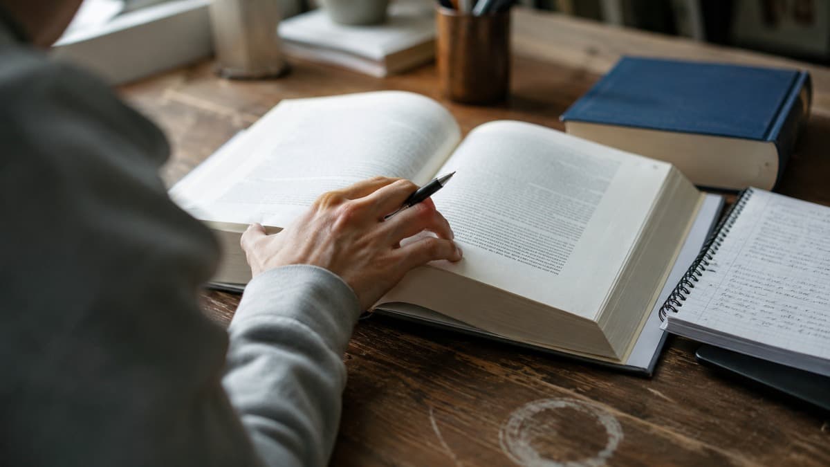 Über die Schulter fotografierte Hand mit Stift auf einem aufgeschlagenen dicken Buch, daneben ein Notizblock mit Handschrift auf einem Holztisch