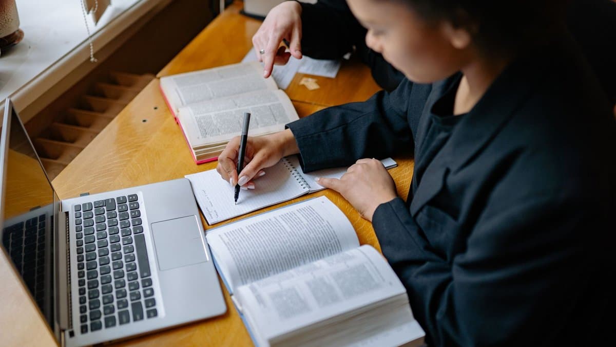 Studentin recherchiert am Laptop mit aufgeschlagenen Büchern und Notizen an einem Holztisch