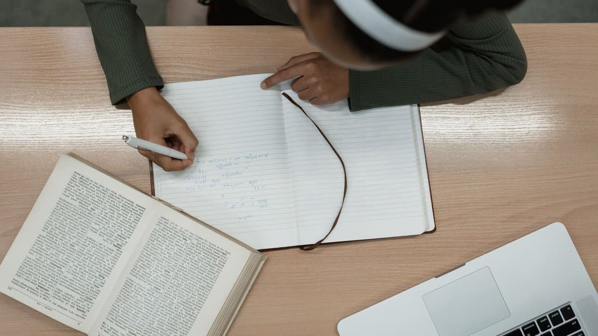 Blick von oben auf einen Schreibtisch mit aufgeschlagenem Buch, handschriftlichen Notizen und einem Laptop