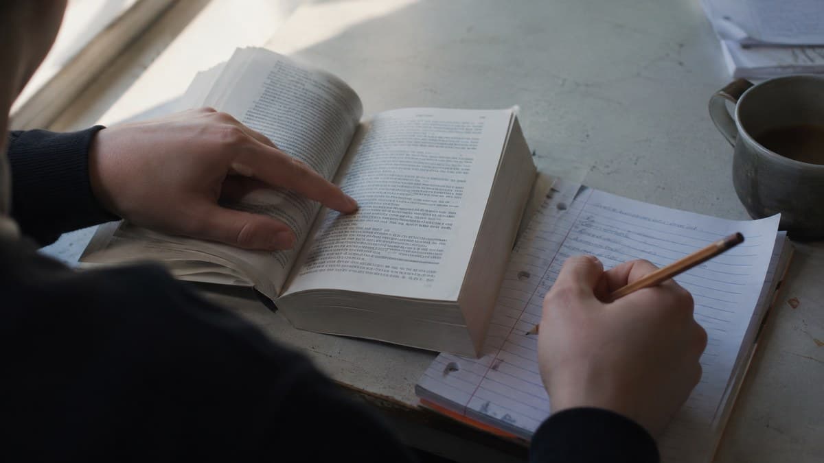 Jemand zeigt mit dem Finger auf eine Zeile in einem aufgeschlagenen Buch und schreibt mit der anderen Hand Notizen auf ein liniertes Blatt