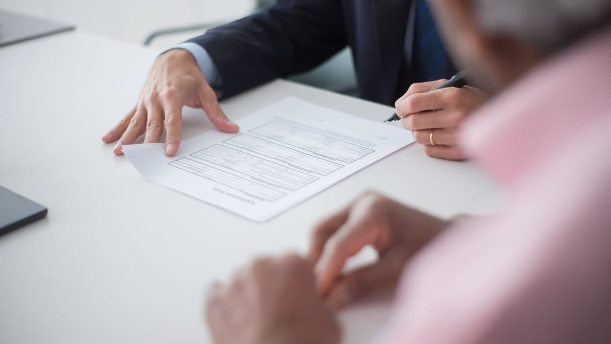 Zwei Personen am Tisch, ein Antragsformular wird über die Tischplatte geschoben, eine Hand hält einen Stift