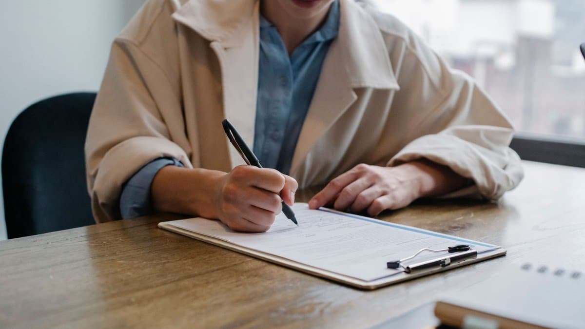 Junge Frau sitzt am Holztisch und füllt handschriftlich ein Formular auf einem Klemmbrett aus