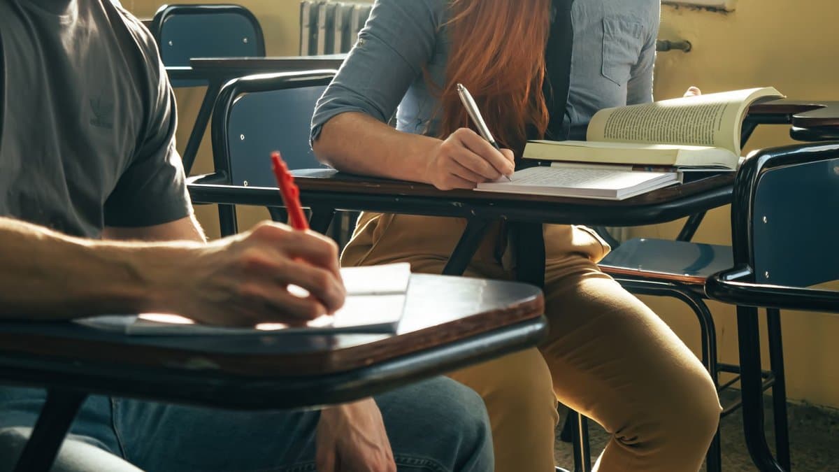 Zwei Studierende schreiben handschriftlich an Klapptischen in einem Seminarraum, neben ihnen liegt ein aufgeschlagenes Buch