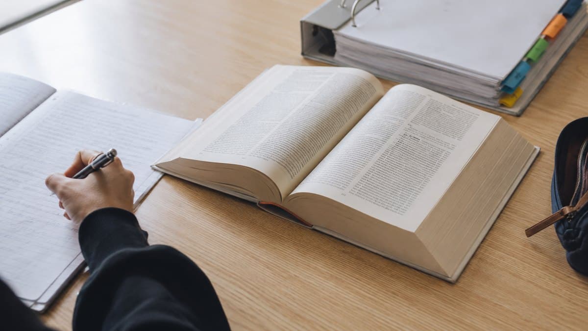 Aufgeschlagenes Gesetzbuch auf einem Schreibtisch, daneben eine Hand, die handschriftlich auf liniertem Papier schreibt, dahinter ein Ordner mit Registerreitern
