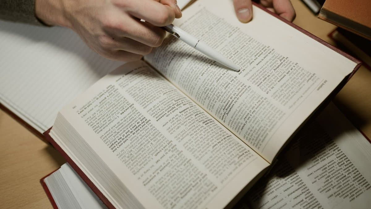 Hand mit Stift zeigt auf eine Textstelle in einem aufgeschlagenen Kommentarband, daneben weitere Bücher und ein Notizblock