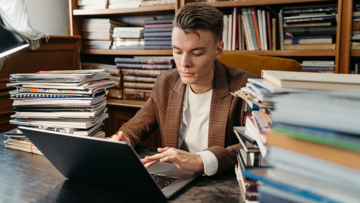 Junger Mann am Bibliothekstisch zwischen Bücherstapeln und Zeitschriften, schreibt am Laptop