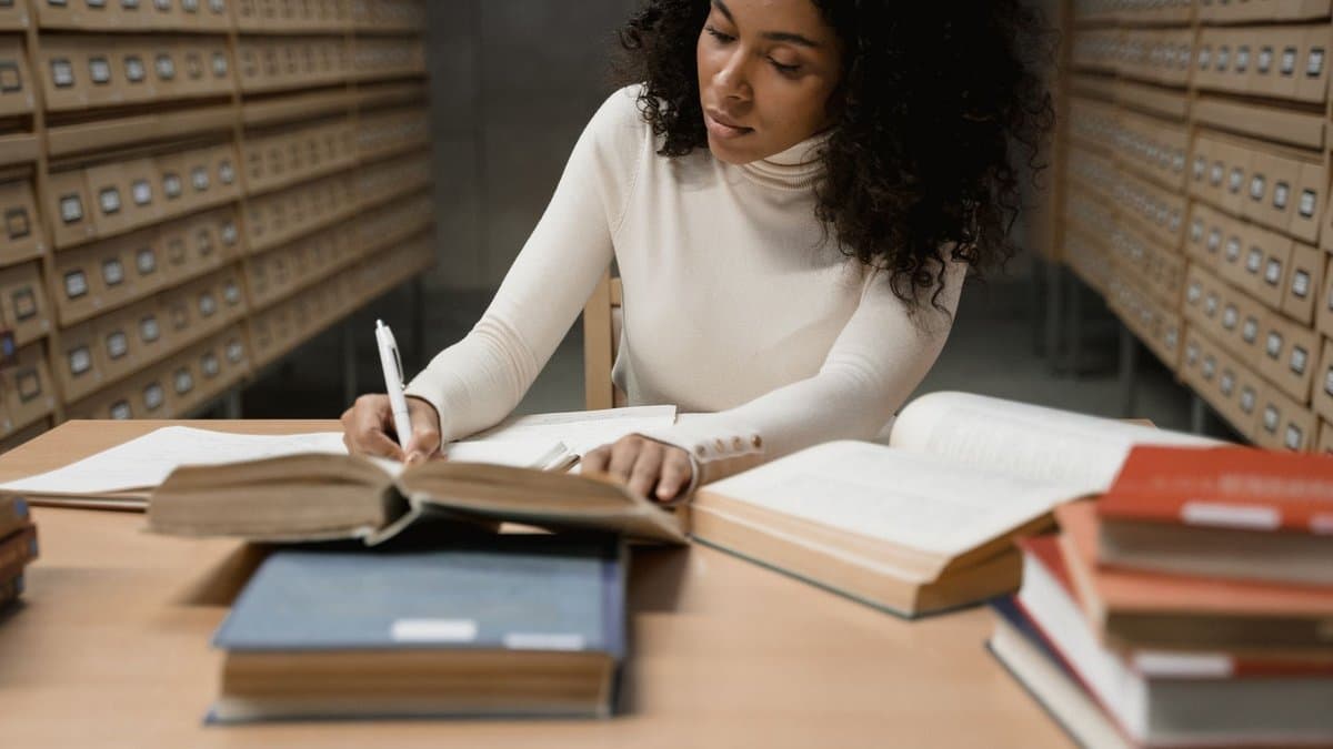 Studentin sitzt in einer Bibliothek zwischen aufgeschlagenen Büchern und schreibt Notizen auf Papier