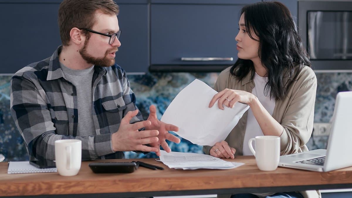 Zwei Menschen sitzen am Küchentisch, eine Frau hält ein Schreiben hoch, ein Mann erklärt ihr etwas mit den Händen