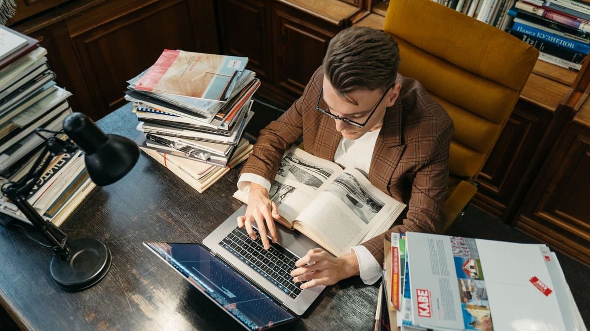 Junger Mann sitzt in einer Bibliothek am Schreibtisch, arbeitet am Laptop und blättert dabei in einem aufgeschlagenen Buch