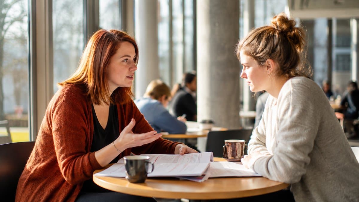 Zwei Studentinnen sitzen an einem runden Tisch in einem hellen Uni-Foyer und besprechen eine korrigierte Klausur