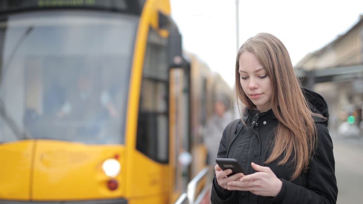 Junge Frau schaut an einer Haltestelle vor einer gelben Straßenbahn auf ihr Smartphone