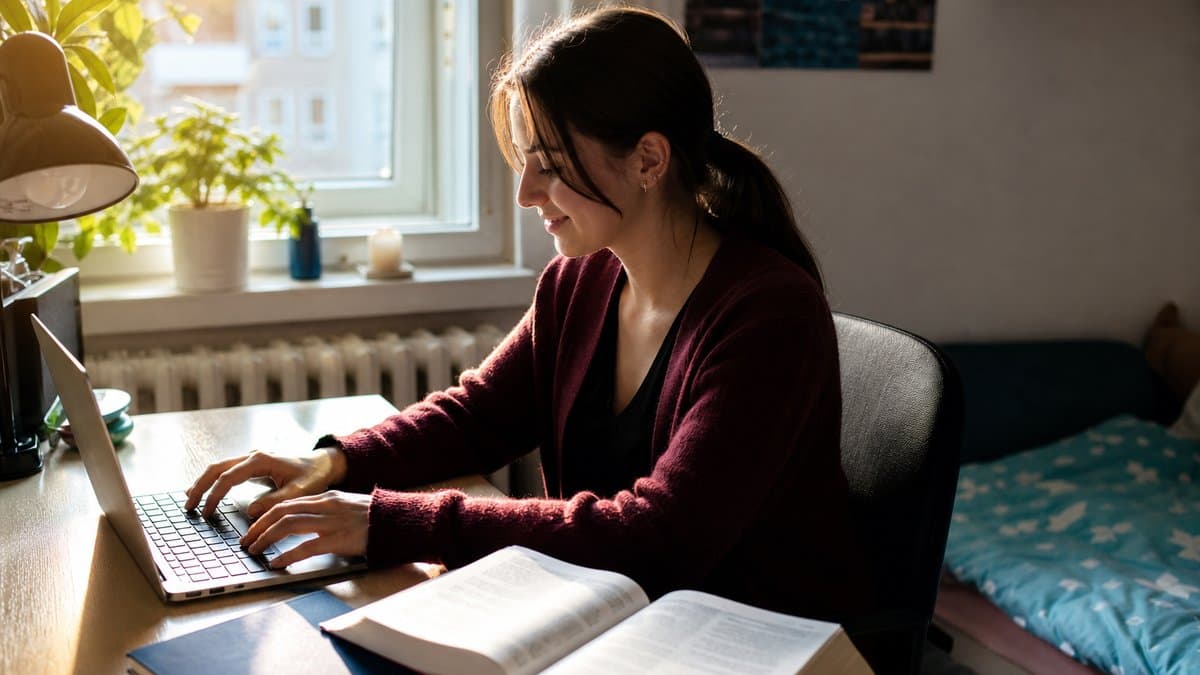 Studentin sitzt morgens am Schreibtisch und tippt am Laptop, daneben liegt ein aufgeschlagenes Lehrbuch unberührt