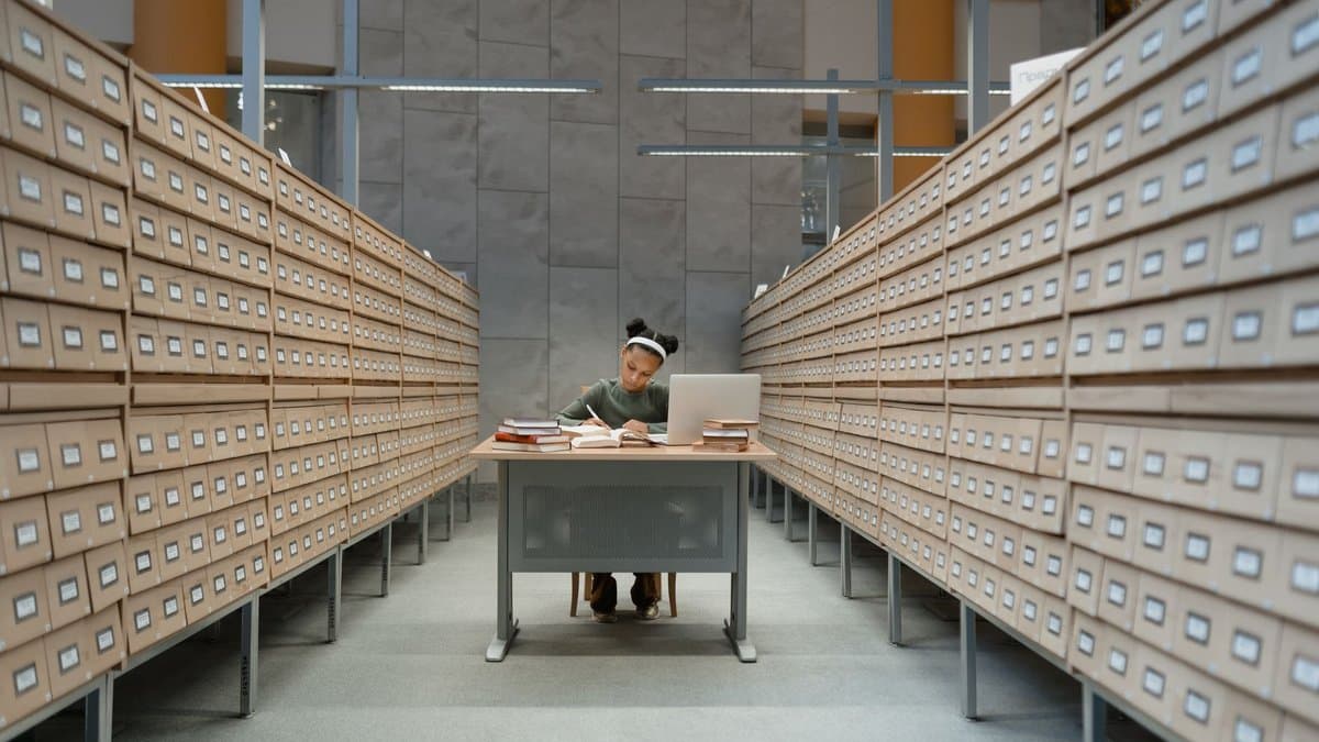 Studentin sitzt an einem Tisch mitten in einem Archivgang zwischen zwei hohen Wänden aus Karteikästen