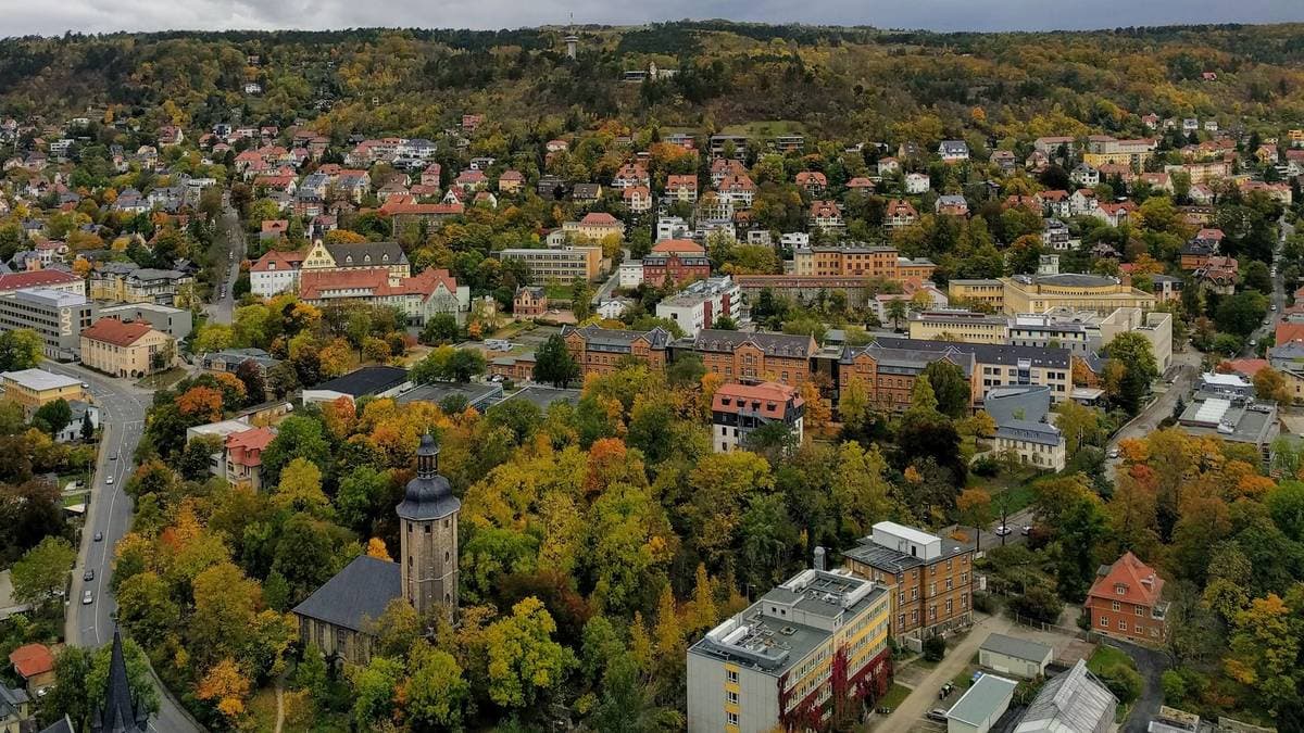 Blick über Jena im Herbst mit Wohnhäusern, Kirchturm und den bewaldeten Hängen des Saaletals