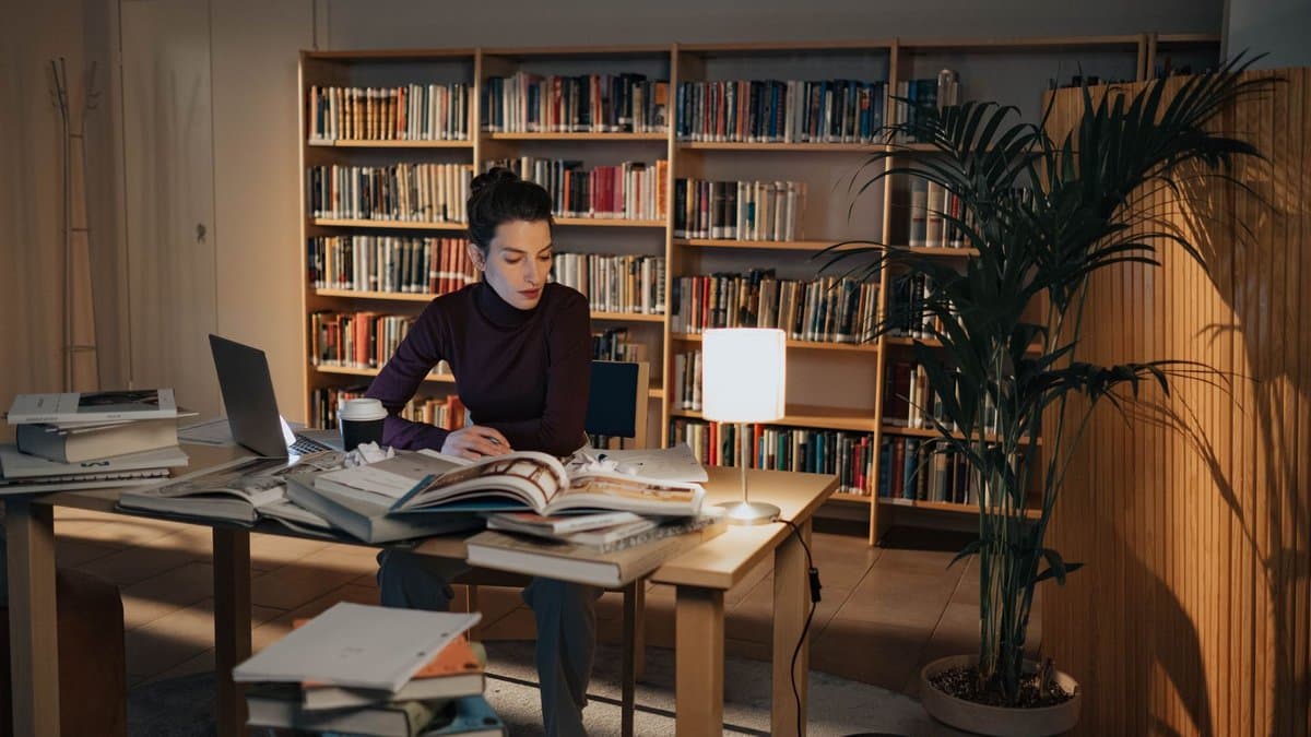 Studentin sitzt abends bei Lampenlicht am Schreibtisch, umgeben von aufgeschlagenen Büchern und Bücherstapeln