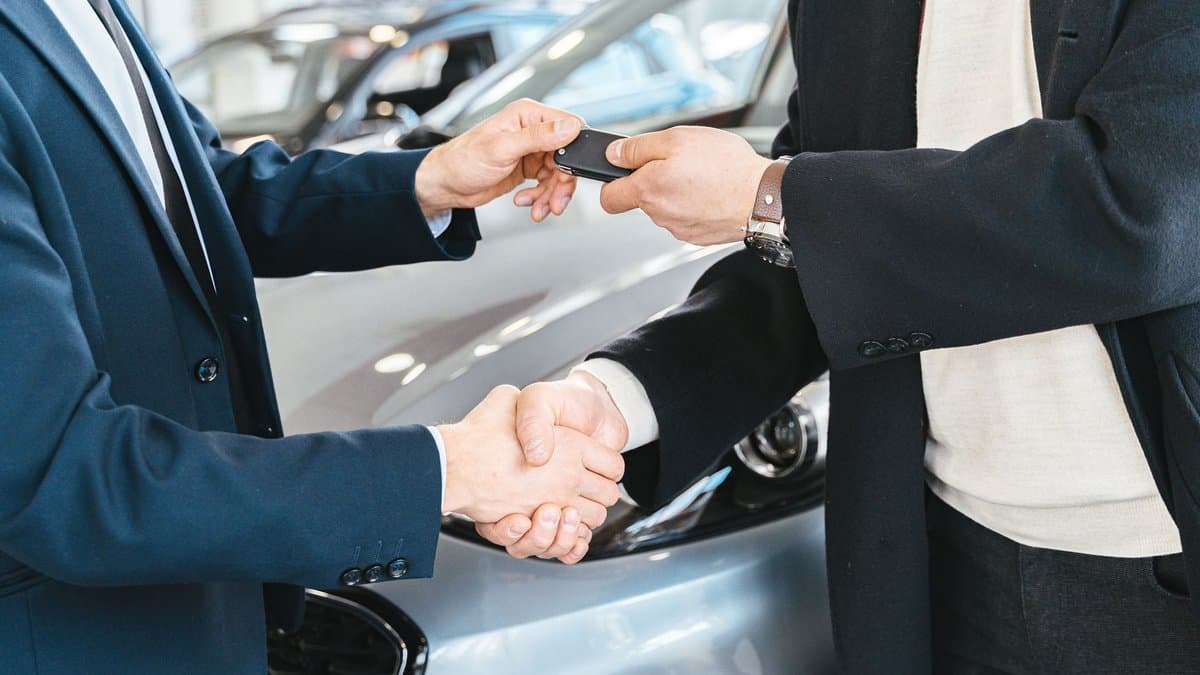Autohändler und Käufer geben sich die Hand und übergeben dabei einen Autoschlüssel vor einem Fahrzeug im Autohaus
