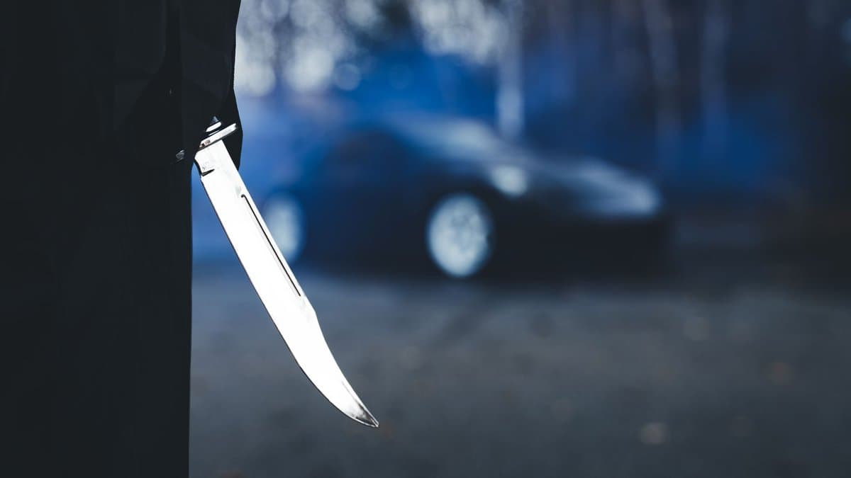 Dunkel gekleidete Person hält ein langes Messer nach unten, im unscharfen Hintergrund ein Auto auf einer Straße in der Dämmerung