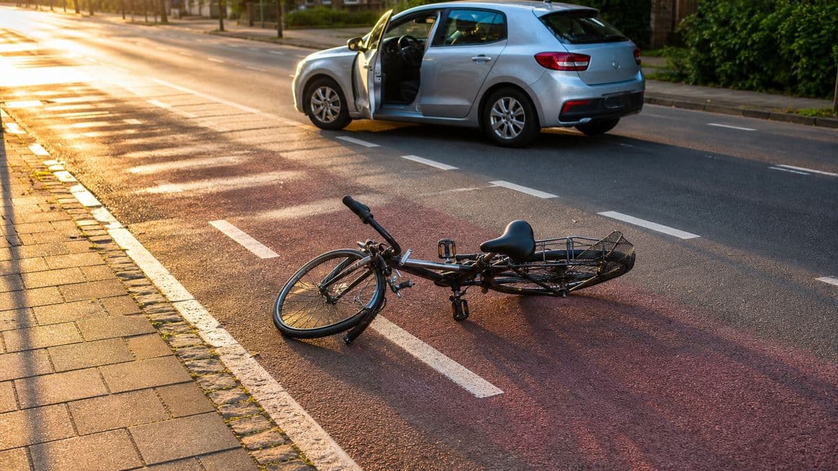 Umgekipptes Fahrrad mit verbogenem Vorderrad auf einem Radweg, dahinter ein angehaltenes Auto mit offener Fahrertür