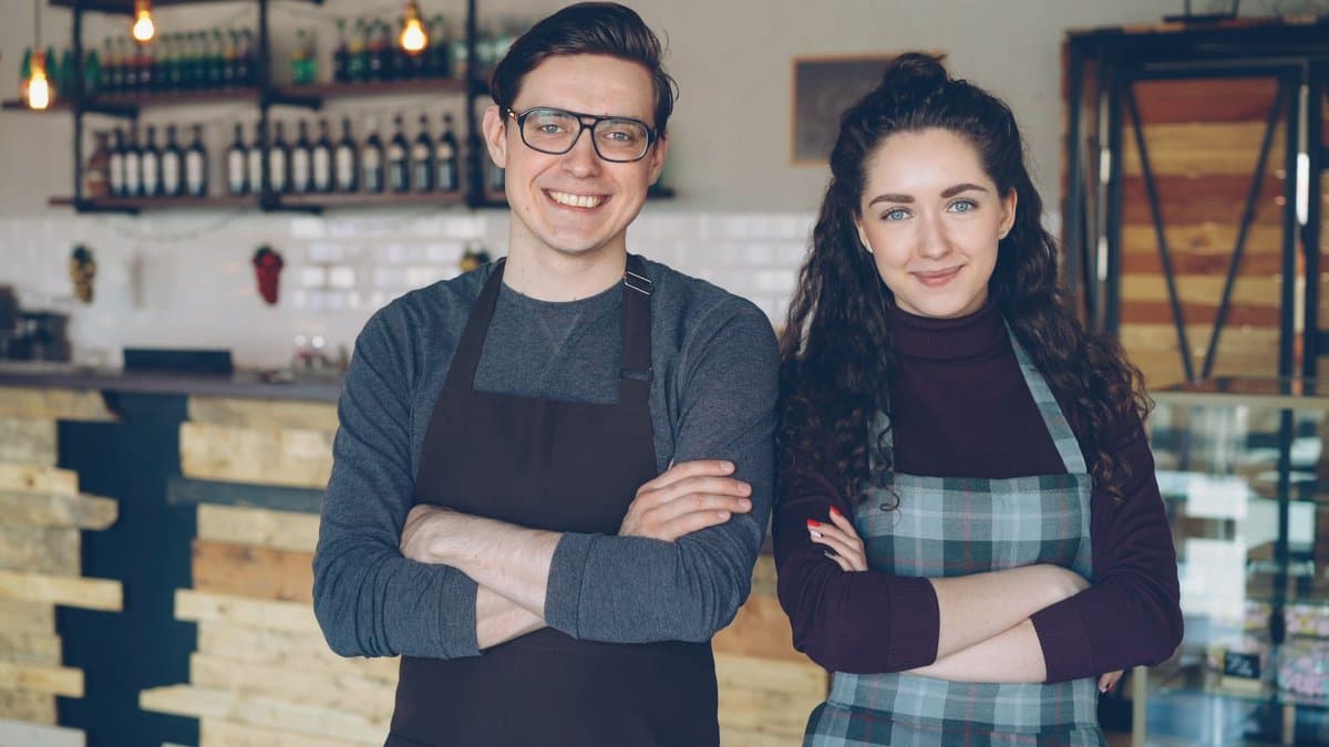Zwei junge Café-Betreiber mit Schürzen stehen nebeneinander in ihrem gemeinsamen Lokal